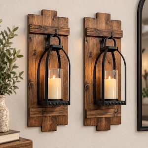 May include: Two wall-mounted candle sconces featuring rectangular dark brown wood planks with attached black metal lantern frames. Each lantern holds a clear glass cylinder containing a white pillar candle with a warm glowing flame.
