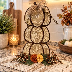 May include: A vertical stack of three pumpkin-shaped cutouts made of dark wood with beige fabric inserts featuring a damask pattern. The top is adorned with a jute twine bow and a wire tendril. The base is decorated with orange and yellow flowers.