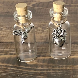 May include: Two small glass bottles with cork stoppers and silver charms. The bottle on the left has a hummingbird charm, and the bottle on the right has a heart charm with floral accents.