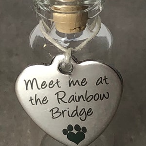 May include: A small glass bottle with a cork stopper and a silver heart-shaped charm attached. The charm has the text "Meet me at the Rainbow Bridge" and a paw print.