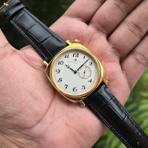 Op de afbeelding: Een goudkleurig polshorloge met een witte wijzerplaat, zwarte Arabische cijfers en een kleine secondewijzer. Het horloge heeft een gouden kast en een zwarte leren band met een alligatorpatroon. Op de wijzerplaat staat de tekst Vacheron Constantin Geneve.