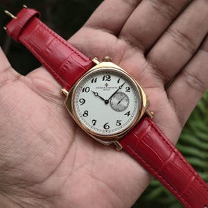 May include: A gold-tone wristwatch with a white dial, black Arabic numerals, and a small seconds sub-dial. The watch features a red leather strap with a textured crocodile pattern and the text Vacheron Constantin Geneve printed on the dial.