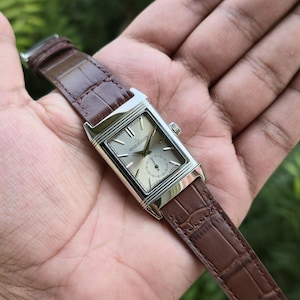 Op de afbeelding: Een rechthoekig zilverkleurig polshorloge met een licht champagnekleurige wijzerplaat, zilveren indexen en een kleine secondewijzer. Op de wijzerplaat staat de tekst Jaeger-LeCoultre. Het horloge heeft een bruine leren band met krokodillenprint.