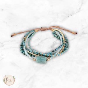Puede incluir: Una pulsera de múltiples hilos con cuentas de color turquesa, cuentas metálicas de color dorado y una cuenta central de piedra rectangular. La pulsera incluye cordones ajustables de color marrón en los extremos.