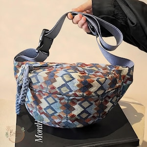 May include: A crescent-shaped crossbody bag featuring a multicolored geometric pattern of blue, white, and rust-orange squares. The bag includes a wide, adjustable light blue strap with a black plastic buckle and a braided blue zipper pull.