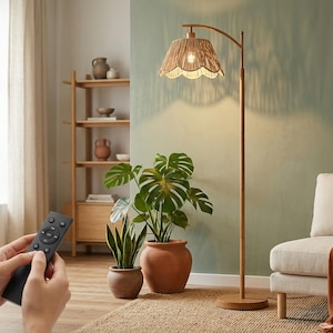 May include: A floor lamp with a tall, thin wooden stand and a round base. The lamp features a scalloped, woven rattan lampshade that emits a warm glow. A black remote control is held in the foreground to operate the lighting fixture.