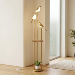 May include: A tall gold floor lamp featuring three vertical poles of varying heights, each topped with a glowing bird-shaped light fixture. The structure includes a circular shelf in the middle and a circular base holding a small potted plant.