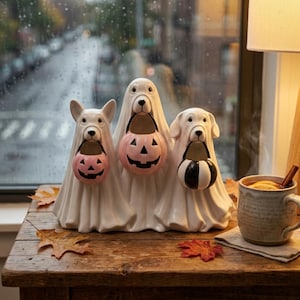 Peut inclure: Une figurine en céramique représentant trois chiens blancs fantomatiques côte à côte. Chaque chien tient un petit seau à friandises dans sa gueule : deux sont des citrouilles roses avec des visages noirs, et un est un seau rayé noir et blanc.