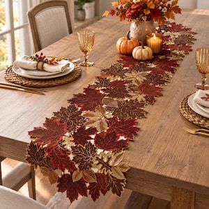 May include: A long table runner featuring an intricate pattern of embroidered autumn leaves in shades of deep red, burgundy, brown, and metallic gold. The runner is laid out on a wooden table, decorated with pumpkins, a candle, and place settings.