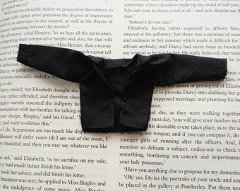 May include: A miniature black jacket with long sleeves, possibly for a doll or small figure, lies on a book page. The jacket has a classic collar and button details, suggesting formal wear.