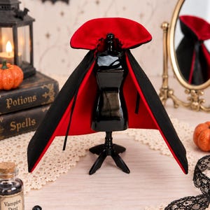 May include: A black cape with a vibrant red lining and a high, stiffened collar displayed on a small black mannequin stand. The cape features thin black ties at the neck and is designed with a wide, flared silhouette that drapes over the shoulders.
