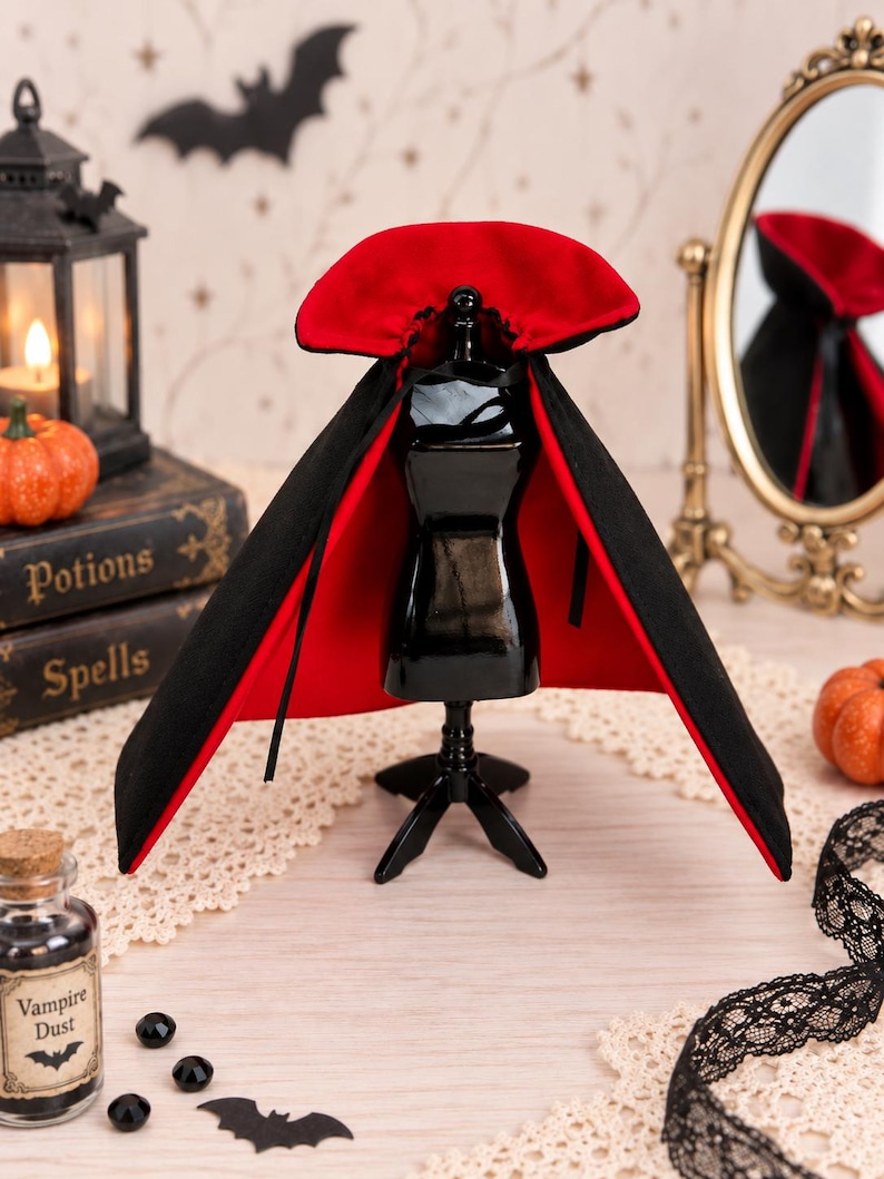 May include: A black cape with a vibrant red lining and a high, stiffened collar displayed on a small black mannequin stand. The cape features thin black ties at the neck and is designed with a wide, flared silhouette that drapes over the shoulders.