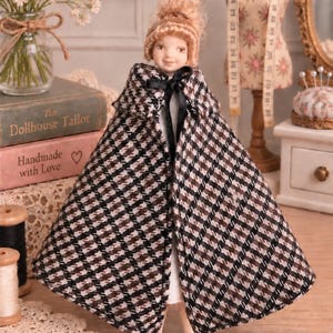 May include: A doll with blonde hair styled in a bun, wearing a brown, black, and white houndstooth patterned cape with a black ribbon tie at the neck. The doll is dressed in a simple white garment underneath the cape and black shoes.