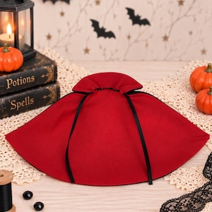 May include: A deep red cape featuring a high, gathered collar and thin black ribbon ties. The garment is constructed from a solid-colored fabric and displays a wide, semi-circular silhouette that drapes outward from the neck area.