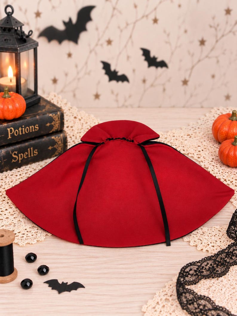 May include: A deep red cape featuring a high, gathered collar and thin black ribbon ties. The garment is constructed from a solid-colored fabric and displays a wide, semi-circular silhouette that drapes outward from the neck area.