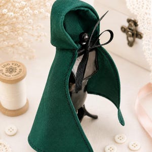 May include: A miniature dark green hooded cloak featuring a black ribbon tie closure at the neck. The garment is draped over a black mannequin form, showcasing a structured hood and a flared hemline with visible black stitching along the edges.
