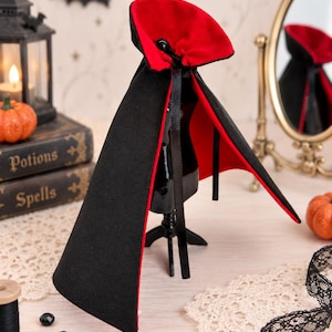 May include: A miniature vampire cape featuring a black exterior and a vibrant red interior lining. The cape includes a high, stiffened collar and thin black ribbon ties at the neck, displayed on a small black mannequin stand for presentation.