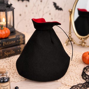 May include: A black drawstring pouch with a vibrant red interior lining. The bag features a wide, rounded base that tapers toward the top opening, which is cinched with a thin black cord. The exterior fabric has a soft, matte texture.