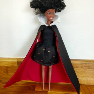 May include: A doll with dark skin and curly black hair wearing a black dress with gold accents and a long black cape featuring a high white collar and a deep red lining. The doll has a neutral expression with purple eyeshadow and a light pink lip color.