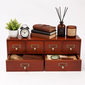 May include: A dark wood desktop organizer with six small upper drawers and two large lower drawers, all featuring brass-toned label pulls. The top surface holds a potted green plant, a black alarm clock, three stacked books, a reed diffuser, and a candle.