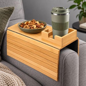 May include: A flexible bamboo sofa arm tray with a slatted design, featuring a built-in square holder for a sage green travel mug and a dark olive bowl filled with mixed nuts, resting securely over the arm of a grey upholstered couch.