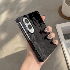 May include: A black smartphone case with a glossy, rippled, liquid-like texture. The case features a raised, oval-shaped camera cutout with two circular lenses and a small flash, showcasing a sleek, reflective surface that mimics flowing water.