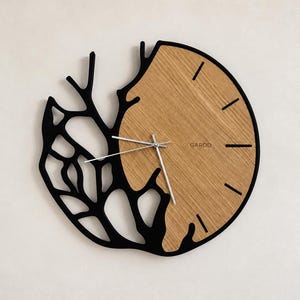 Peut inclure: Une horloge murale circulaire au design divisé. Une moitié est en bois brun clair avec des marqueurs d'heure noirs et le texte GARDD. L'autre moitié est un cadre noir avec un motif découpé complexe en forme de branches. Trois aiguilles argentées.
