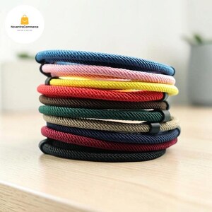 Puede incluir: Una pila de diez pulseras circulares hechas de cordón trenzado en varios colores, incluyendo azul, rosa, amarillo, rojo, marrón, verde, beige, azul marino, granate y negro. Cada pulsera cuenta con una pequeña pieza de conexión rectangular oscura.