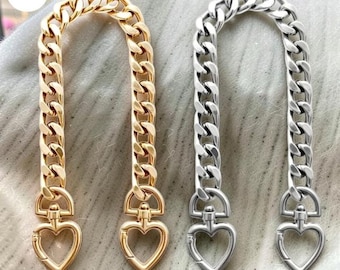May include: Two metal chain straps for bags, one in a gold tone and one in a silver tone. Each strap features thick, interlocking curb links and is finished at both ends with a functional swivel clasp shaped like a heart for secure attachment.