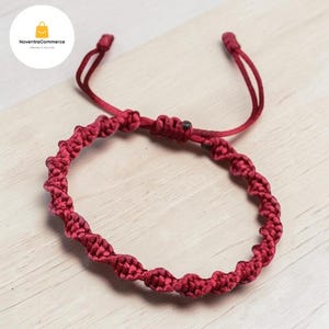 May include: A deep red cord bracelet featuring a continuous series of intricate macrame knots. The bracelet includes an adjustable sliding knot closure with two loose cord ends extending from the central knot mechanism for a secure fit.