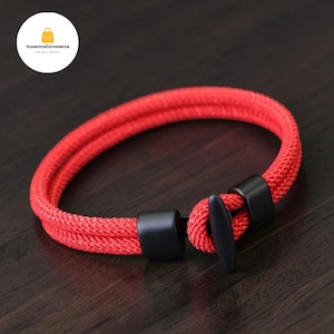 Peut inclure: Un bracelet à double cordon rouge vif doté d'un fermoir à bascule en métal noir mat. Le bracelet est fabriqué à partir d'un matériau synthétique tissé et comprend deux accents en métal noir qui fixent les extrémités du cordon près de la fermeture.