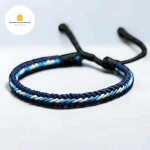 Puede incluir: Una pulsera de cordón trenzado con un patrón de hilos azul oscuro, azul claro y blanco. La pulsera incluye un mecanismo de cierre ajustable con dos extremos de cordón negro largos que se extienden desde la estructura principal.