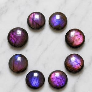 Puede incluir: Ocho piedras preciosas de labradorita circulares y pulidas dispuestas en círculo. Cada piedra presenta una superficie lisa y redondeada con destellos iridiscentes de color púrpura intenso, violeta y azul sobre una base mineral oscura.