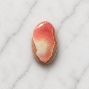 Könnte beinhalten: Ein oval geschliffener Edelstein mit glatter Oberfläche und einem Farbverlauf von tiefem Korallenrosa zu einem cremefarbenen, weißlichen Zentrum. Der Stein zeigt natürliche Mineralbänderungen und eine glänzende, reflektierende Oberfläche.