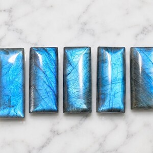 Puede incluir: Cinco piedras preciosas de labradorita rectangulares dispuestas en una fila horizontal. Cada piedra presenta una superficie pulida y lisa con un distintivo destello azul iridiscente y fracturas internas visibles, mostrando un color profundo.