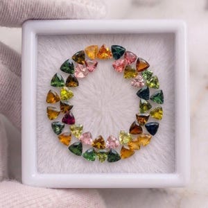 Pode incluir: Uma coleção de pequenas pedras preciosas facetadas com corte triangular dispostas num padrão circular dentro de uma caixa de apresentação quadrada branca. As pedras apresentam uma variedade de cores, incluindo tons de verde, amarelo, laranja, rosa e azul profundo.