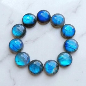 Può includere: Undici cabochon di labradorite rotondi e levigati disposti in un motivo circolare. Ogni pietra mostra un vibrante riflesso blu iridescente sulla sua superficie liscia e bombata, mettendo in risalto fratture interne naturali e toni di base grigio scuro.