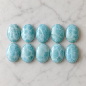 Könnte beinhalten: Zehn ovale Cabochons aus hellblauem Larimar-Stein. Jedes Stück hat eine glatte, polierte Oberfläche mit einzigartigen weißen, wolkenartigen Mustern und verschiedenen Schattierungen von Himmelblau, angeordnet in zwei Reihen zu je fünf.
