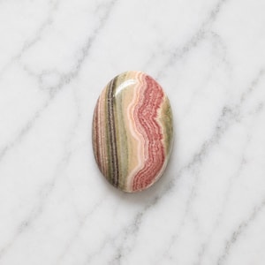 Könnte beinhalten: Ein ovaler Rhodochrosit-Edelstein mit einer polierten, glatten Oberfläche, der deutliche gebänderte Schichten in zartem Rosa, tiefem Pink, cremigem Beige und gedämpftem Olivgrün aufweist und natürliche Mineralmuster sowie ein glänzendes Finish zeigt.