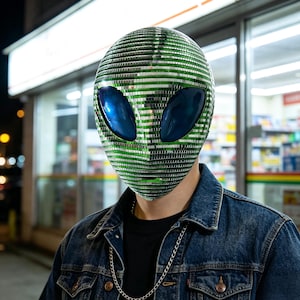 Könnte beinhalten: Eine Alien-Maske für den gesamten Kopf, die mit kleinen, reflektierenden grünen und silbernen Spiegelkacheln bedeckt ist und zwei große, glatte, dunkelblaue ovale Augenlinsen aufweist. Die Maske wird über einem schwarzen Shirt, einer blauen Jeansjacke und einer silbernen Kette getragen.
