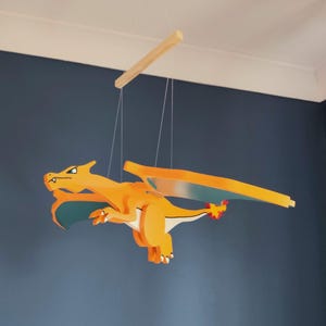 Könnte beinhalten: Ein hängendes Mobile mit einer hölzernen, orangefarbenen Drachenfigur, deren Flügelunterseiten blaugrün gefärbt sind und die eine Flamme an der Schwanzspitze trägt. Die Figur hängt an dünnen weißen Schnüren an einer horizontalen Holzstange.