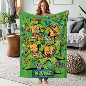 May include: A green fleece throw blanket featuring a graphic print of four Teenage Mutant Ninja Turtles characters in action poses with their signature weapons, pizza slices, and ninja stars. The bottom includes a rectangular box labeled NAME.