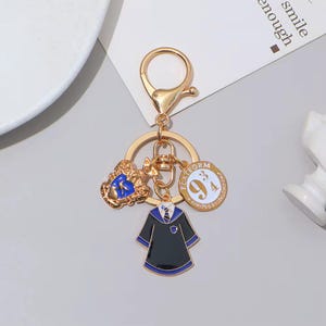 May include: A gold-toned metal keyring featuring three charms: a blue and gold crest, a circular charm with the text "PLATFORM 9 3/4 KING'S CROSS LONDON" in white and gold, and a black and blue robe charm with a striped tie and collar detail.