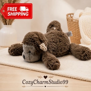 May include: A plush monkey toy with dark brown shaggy fur and a smooth grey face. The stuffed animal is in a lying position with a curled tail and small light-colored ear details. A small white tag is visible on the side of the monkey's back.