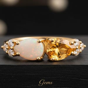 May include: A gold ring featuring a pear-shaped white opal and a pear-shaped yellow citrine gemstone side by side. The band is accented with small, clear round stones on both sides of the central gems. The word Gems is printed below the ring.