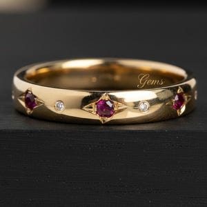 Peut inclure: Une bague en or ornée de rubis rouges ronds et de petits diamants transparents alternés incrustés dans le métal. Les rubis sont sertis dans des gravures en forme d'étoile, tandis que les diamants sont dans des chatons ronds. Texte Gems visible.