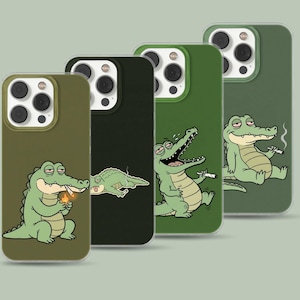 May include: Four smartphone cases in shades of olive and dark green, each featuring a cartoon alligator illustration. The designs show the alligator in various poses, including lighting a cigarette, lying down, laughing, and sitting while holding a cigarette.