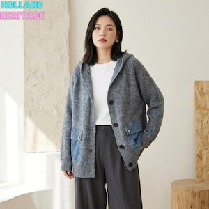 Puede incluir: Un cárdigan de punto gris con capucha, cierre de botones, mangas largas y dos bolsillos de parche de mezclilla azul. La prenda se lleva sobre una camiseta blanca lisa y pantalones gris oscuro. El texto HOLLAND HERITAGE aparece arriba a la izquierda.