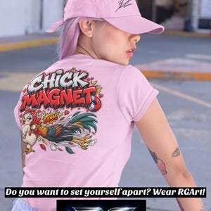 Op de afbeelding: Een lichtroze honkbalpet met een zwart geborduurd logo en een lichtroze t-shirt met een kleurrijke afbeelding op de achterkant. De afbeelding toont een cartoonkip en haan met de tekst CHICK MAGNET en BAMM in een graffitistijl.