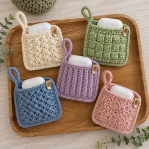 May include: Five square crochet pouches in cream, sage green, lavender, dusty blue, and soft pink, each featuring a unique textured stitch pattern, a small hanging loop, and a white wireless earbud charging case tucked inside with a small brown tag.